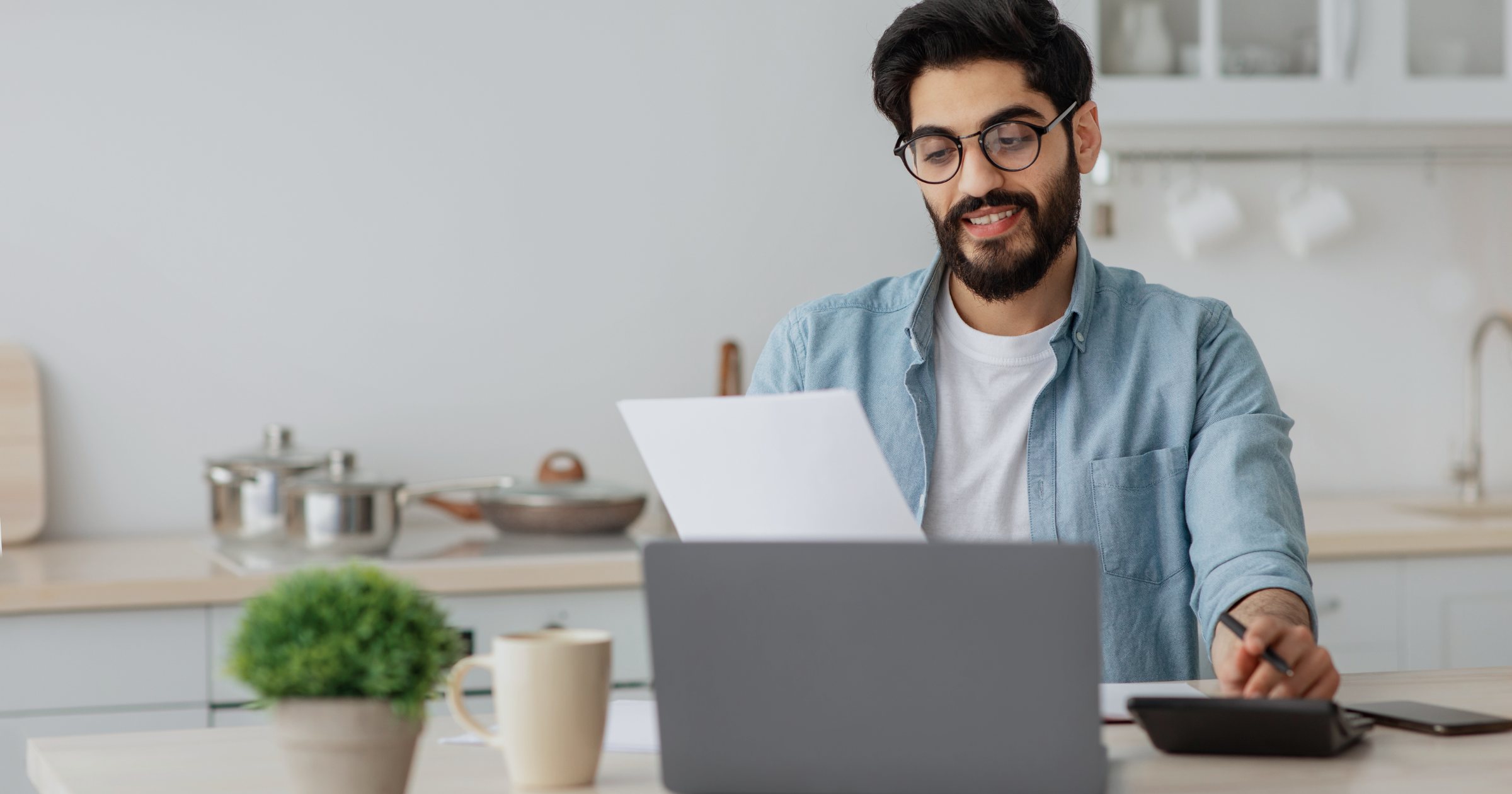 Bărbat care citește atent o ofertă pe hârtie la masa din bucătărie, cu un laptop și un calculator de birou alături