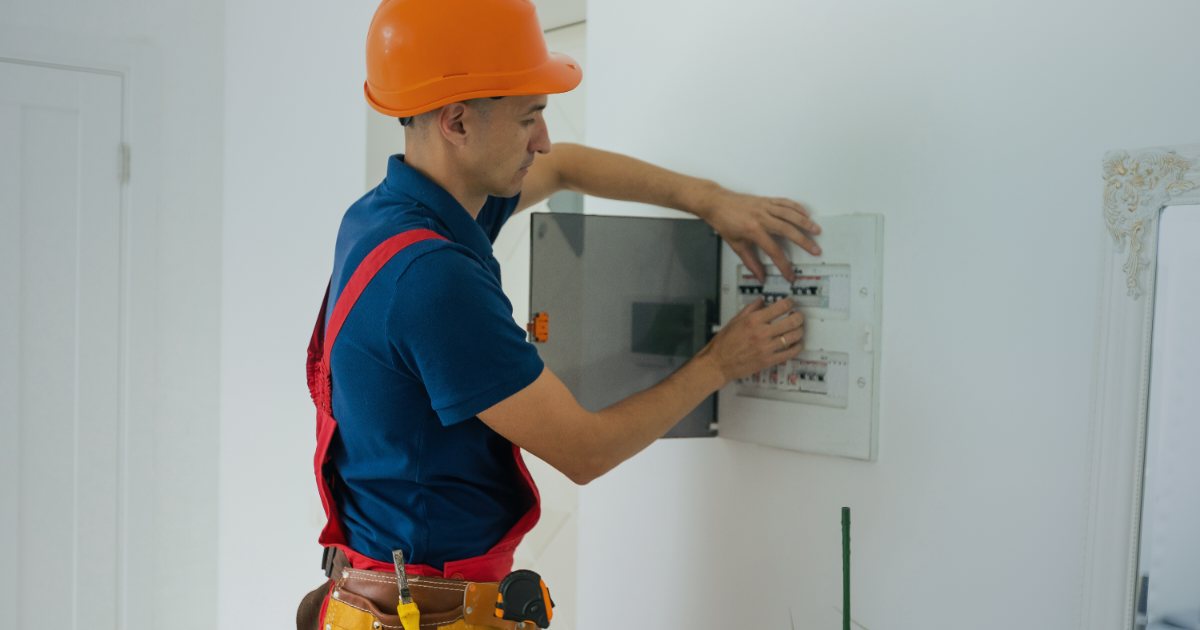 Electrician cu mănuși de protecție lucrând la un tablou electric deschis, verificând siguranțele și cablajul instalației