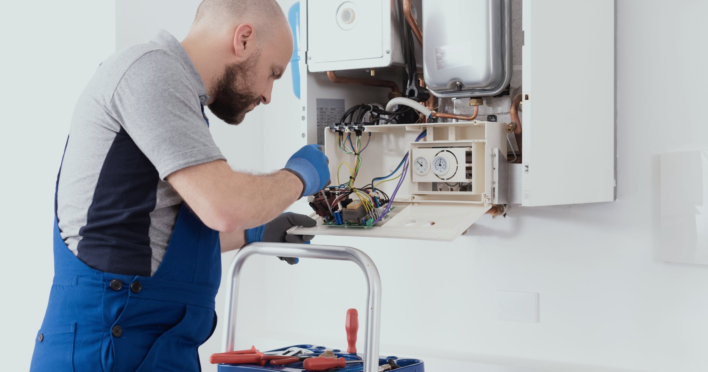 Tehnician în salopetă reparând o centrală termică pe gaz deschisă, cu sculele la îndemână, la fața locului